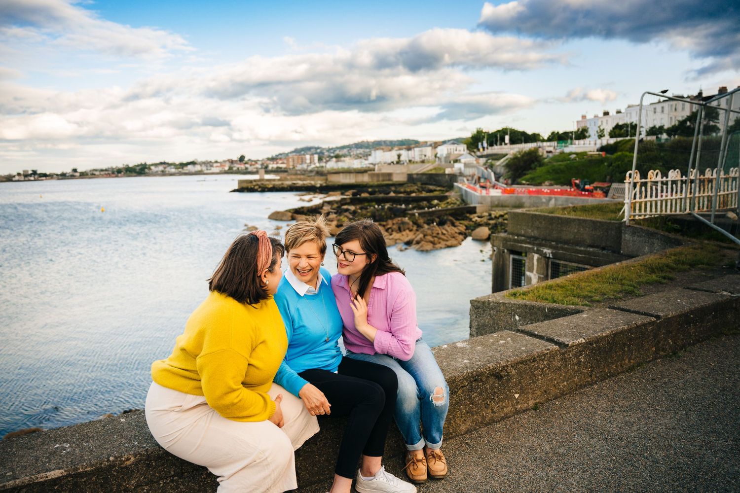 Three people sit together laughing on a waterfront wall with a coastal town and cloudy sky in the background.