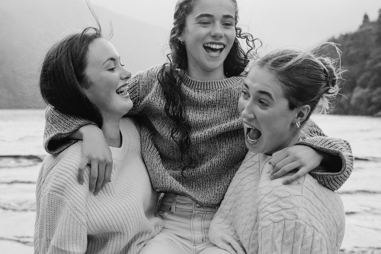 Three friends in cozy sweaters share genuine laughter together during a black and white photoshoot at the beach. family PHOTOGRAPHER Dublin