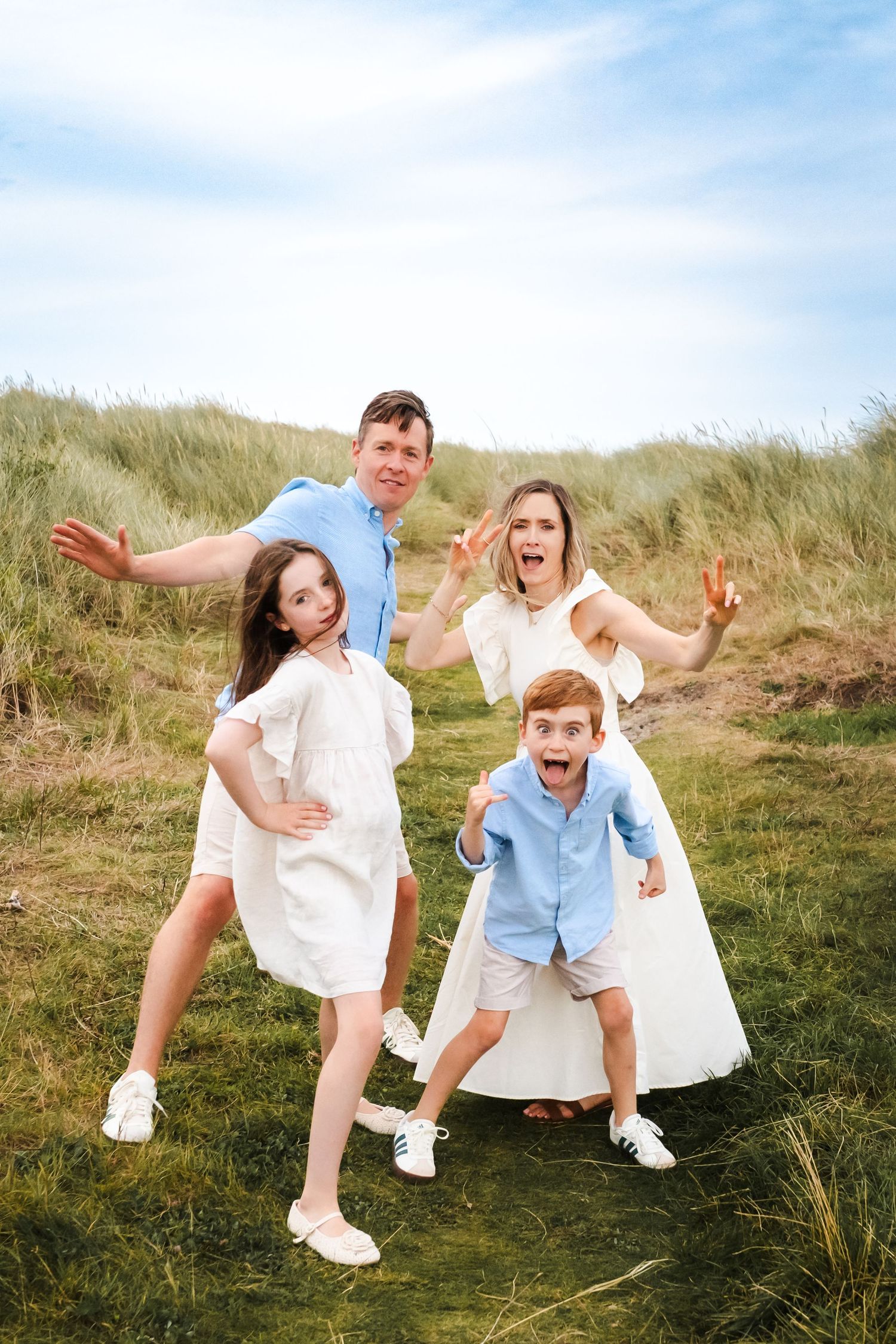 A playful family makes silly poses together in grassy dunes on a sunny day.  Family photographer Dublin Ireland