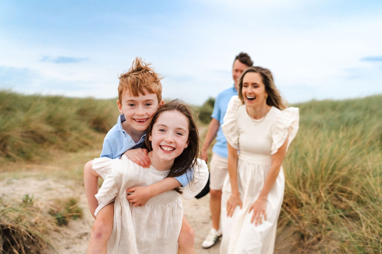 A joyful group plays and laughs together on scenic coastal sand dunes during golden hour.  Family photographer Dublin Ireland