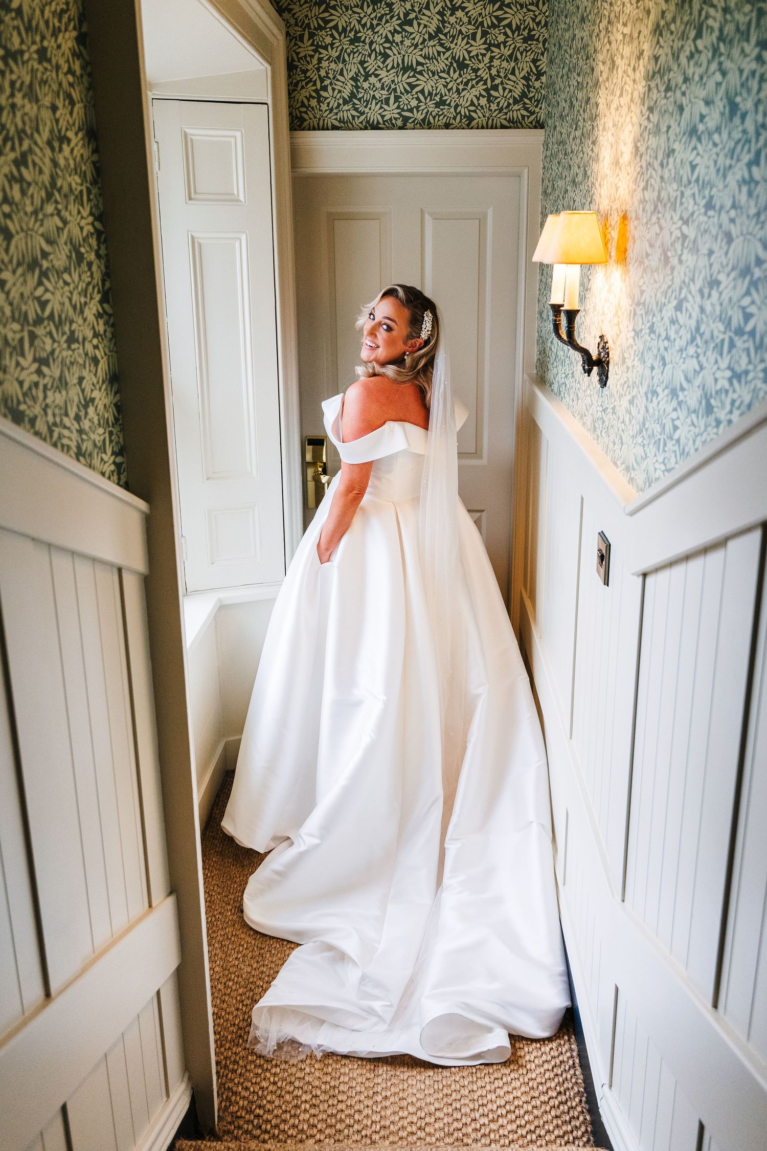 Elegant white wedding gown on display in a room with floral wallpaper and white doors.  Virginia Park Lodge Wedding Photographer Cavan Dubli