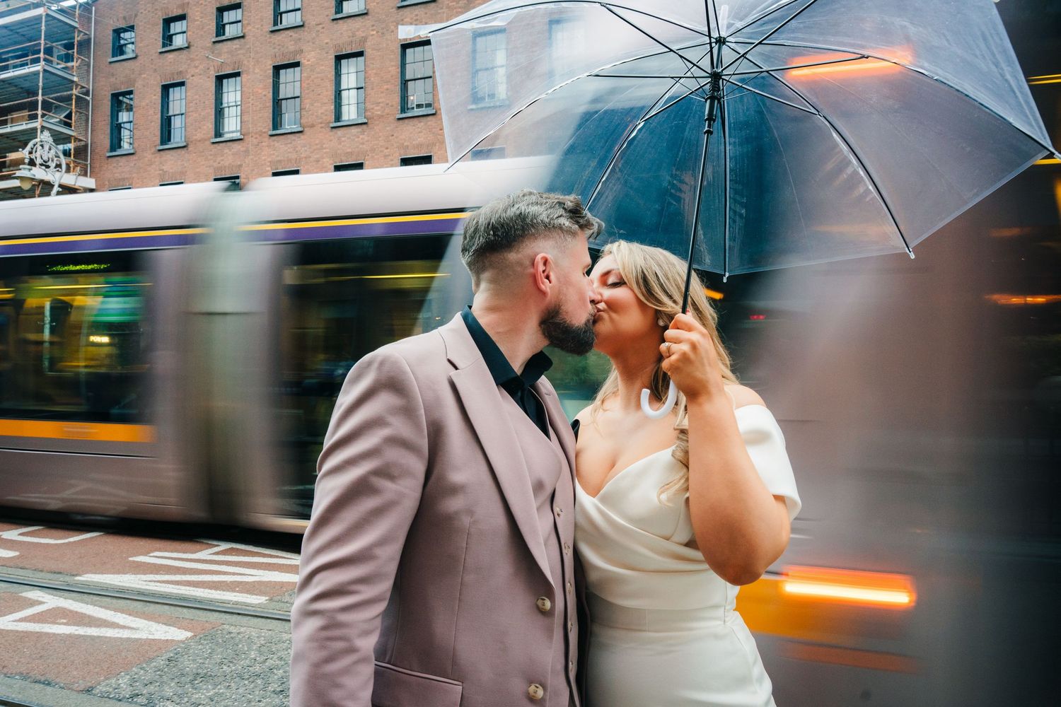 Romantic couple shares a kiss under clear umbrella with motion-blurred city tram passing behind them on rainy street. Wedding PHOTOGRAPHER D