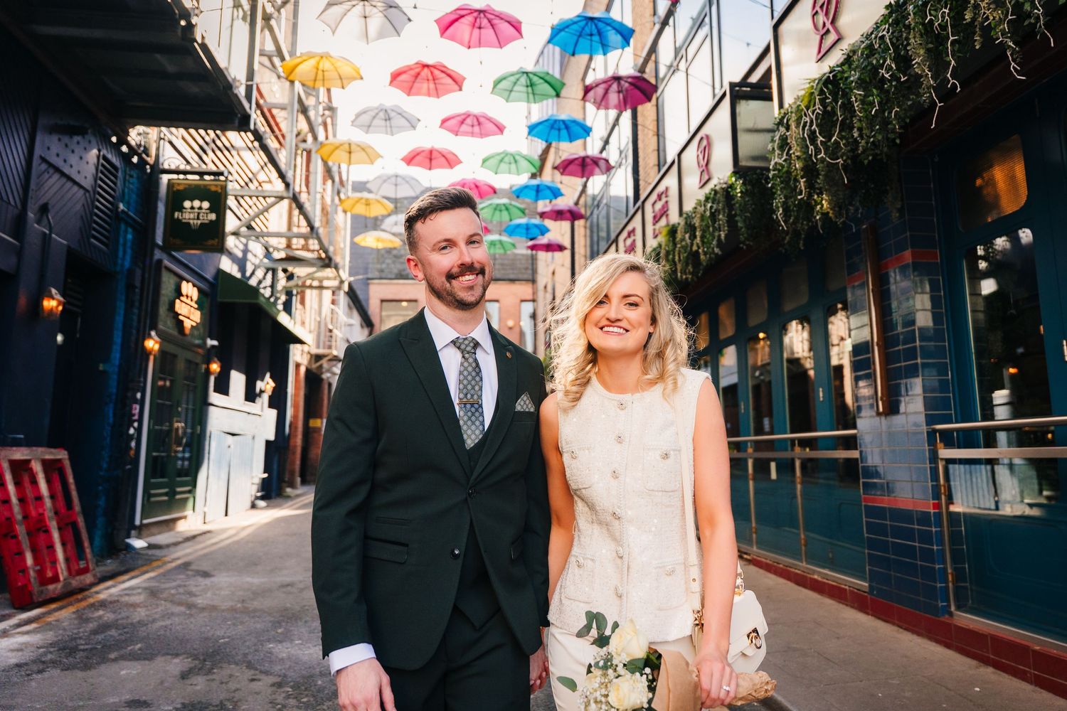 A couple poses under colorful hanging umbrellas in an urban alleyway decorated for celebration. City Hall Dublin wedding photogrAPher