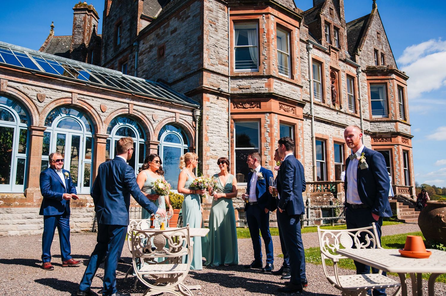 A gathering of people celebrating outdoors in front of a grand Victorian-style mansion with red sandstone exterior and ornate details. Castl