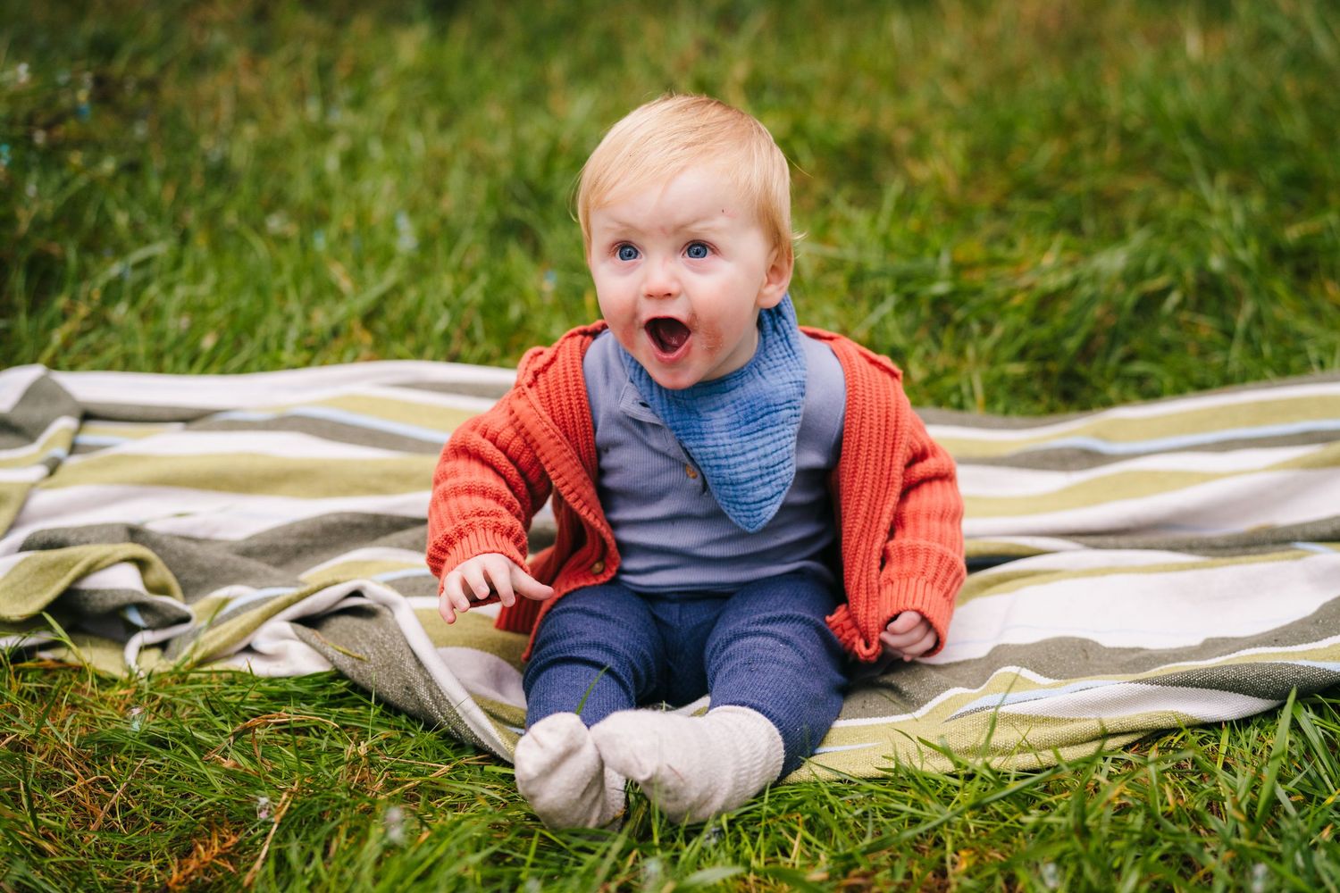 Baby sits on blanket during outdoor photoshoot with expressive reactions and natural lighting.