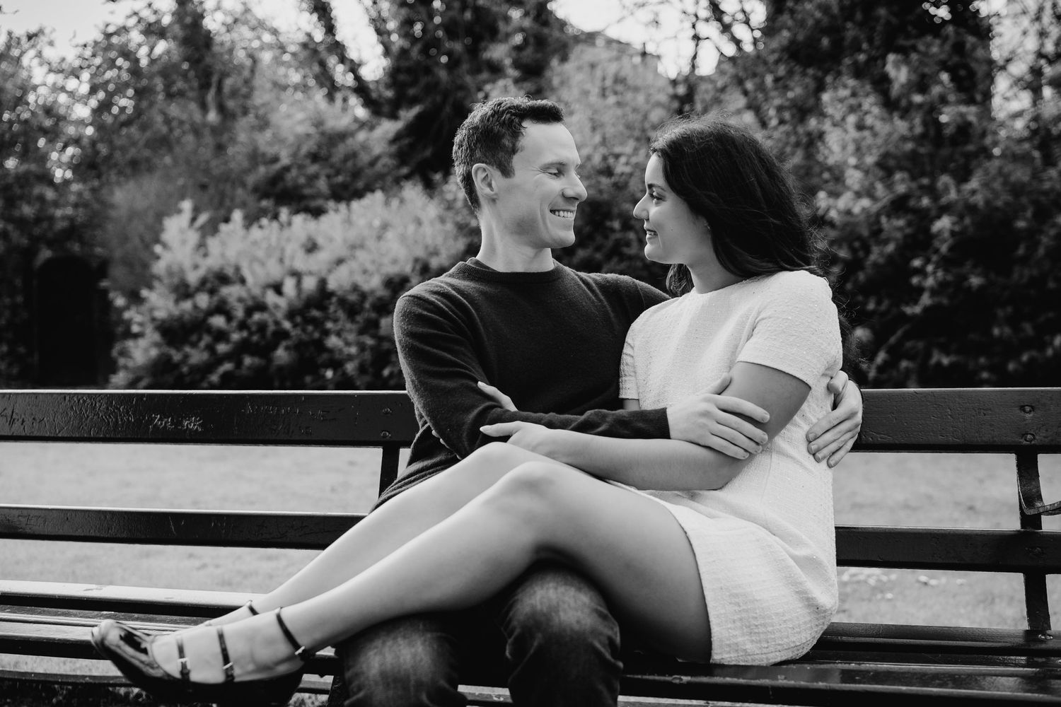 A couple shares an intimate moment while sitting together on a wooden park bench in black and white. Surprise proposal dublin photography