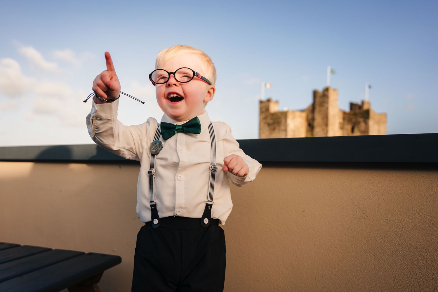 A stylesh boy in suspenders and bowtie runs playfully through a field at golden hour.  Wedding PHOTOGRAPHER Trim Castle