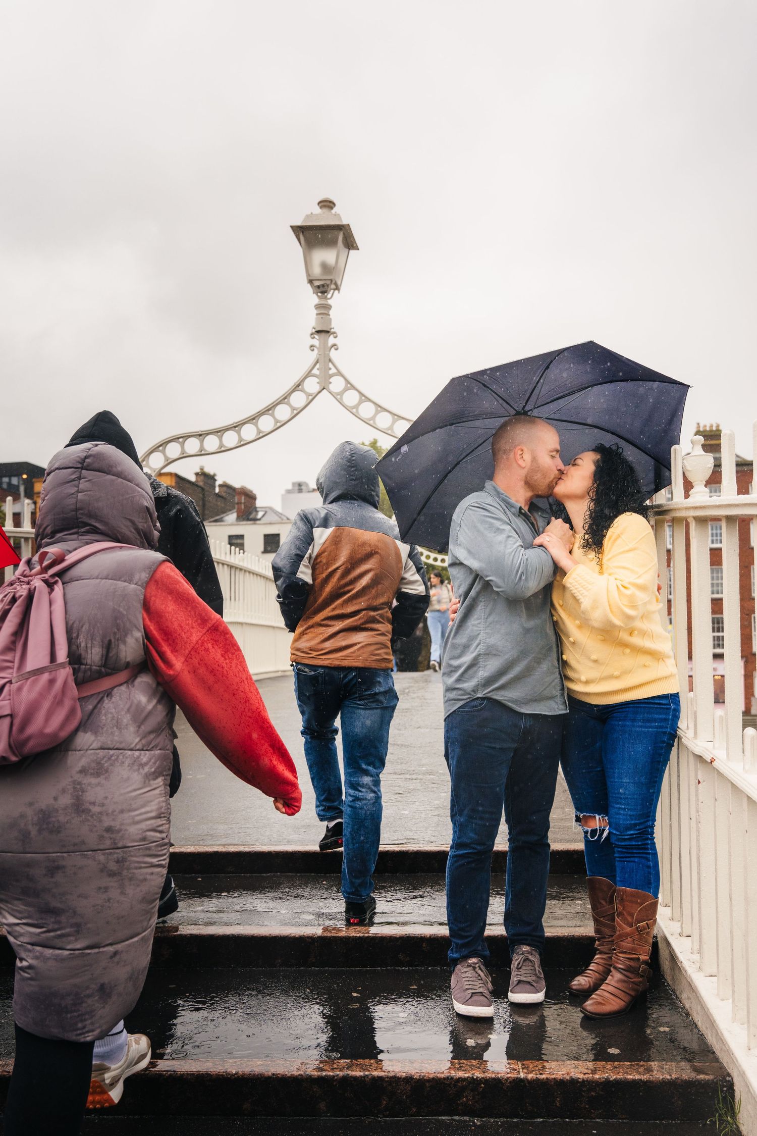 Romantic couple sharing umbrellas while standing along white railings on an overcast day. Proposal Dublin Photographer