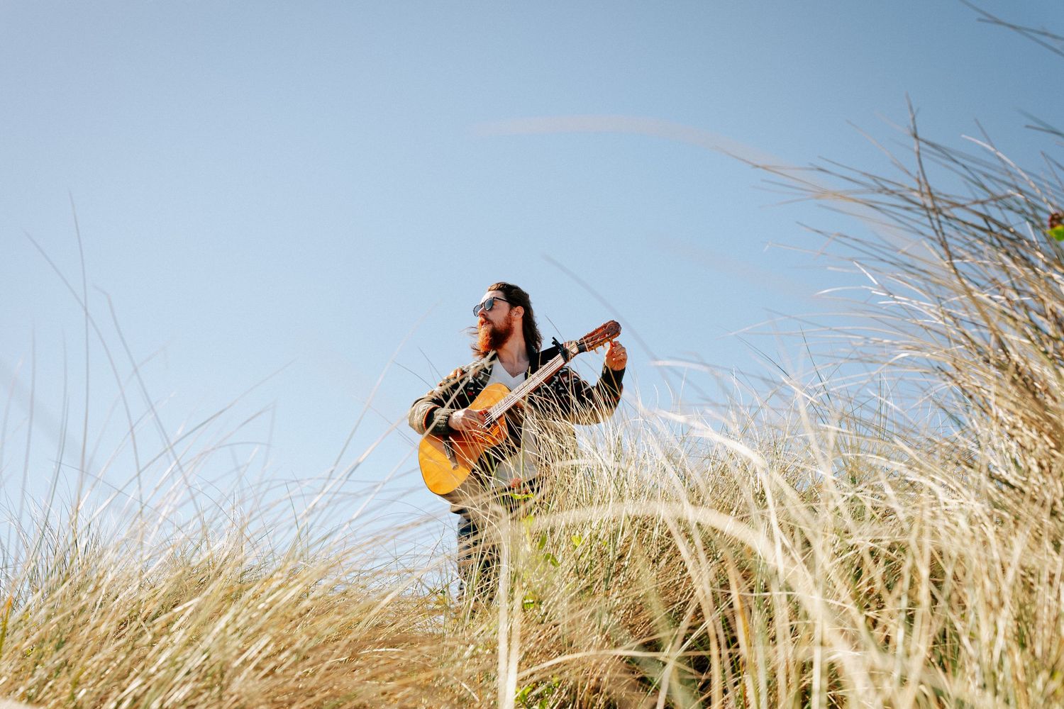 Guitar player sits in tall grass playing acoustic guitar against blue sky backdrop. PHOTOGRAPHER Dublin