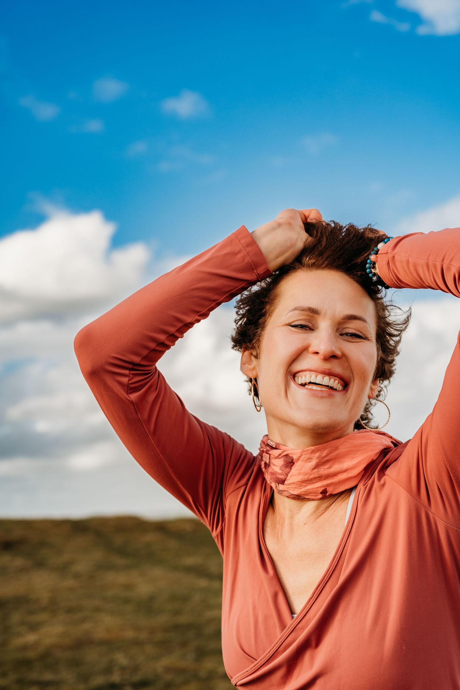 Person wearing coral-colored top smiles joyfully with raised arms against blue sky and rolling hills backdrop. Portrait PHOTOGRAPHER Dublin