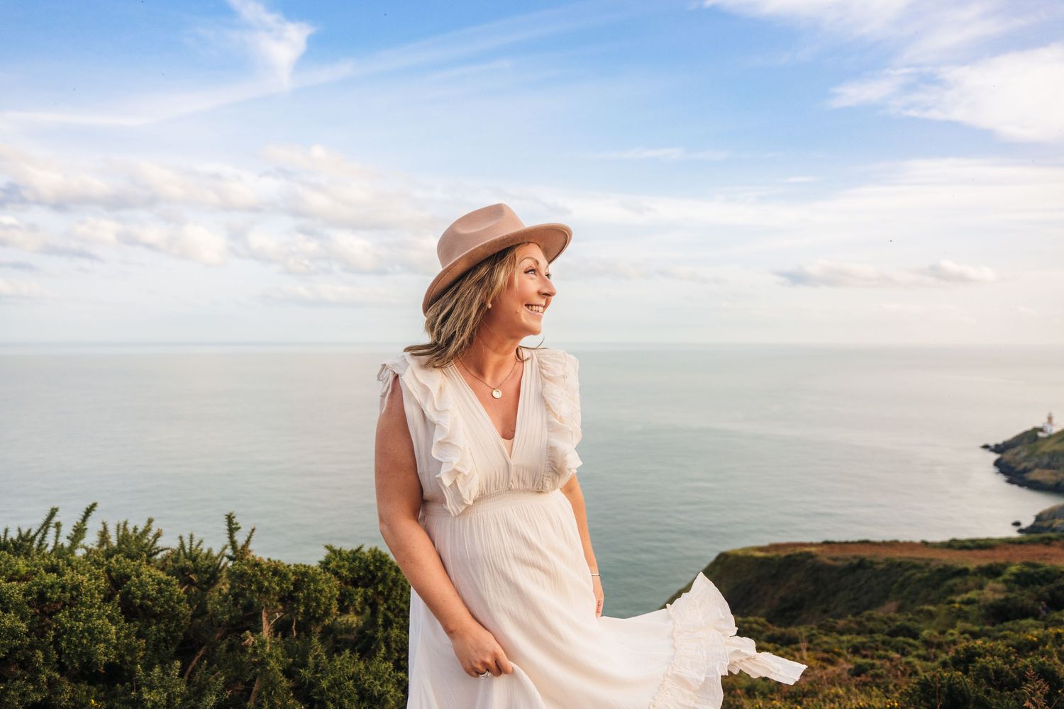 A scenic coastal view with a person in a white dress walking along the cliffside against a bright blue sky. Family photographer dublin i