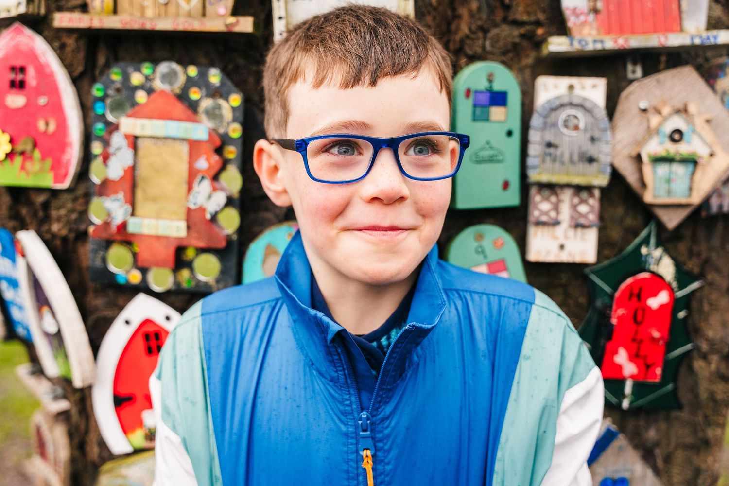 A person wearing glasses and blue clothing sits in front of a colorful wall covered in artwork and decorative items. Family photographer Dub