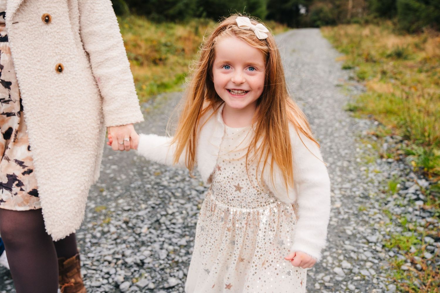 A young child in a sparkly cream dress walks along a gravel path while holding an adult's hand. Family Photographer Dublin Ireland