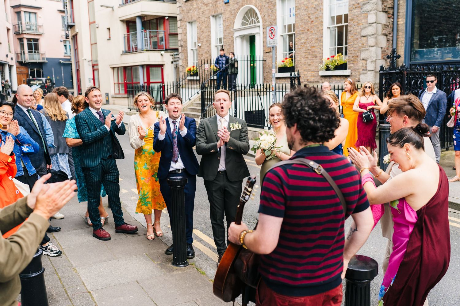 Wedding guests applaud and celebrate outside a historic brick building. PHOTOGRAPHER Dublin Ireland Wedding