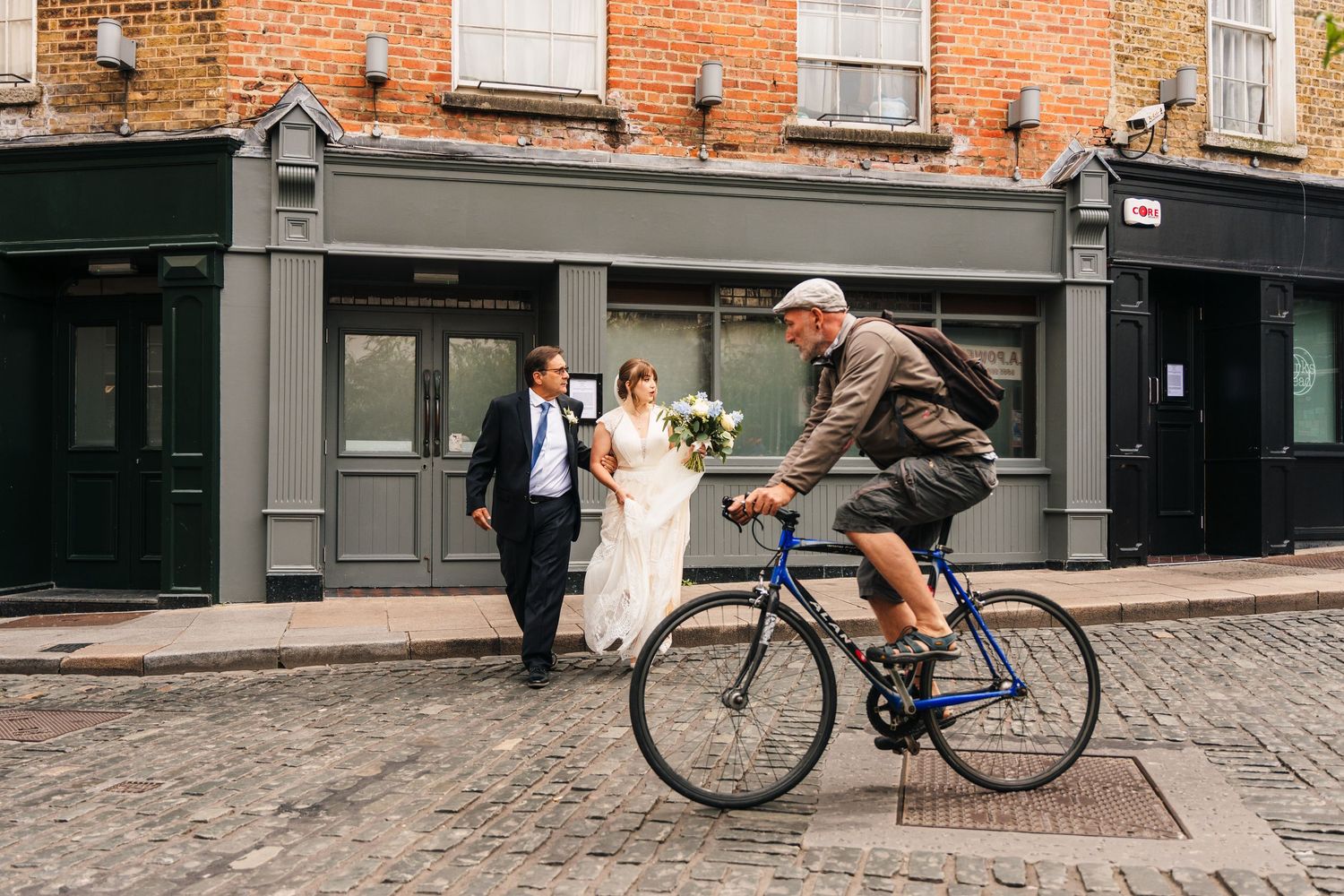 A cyclist rides past a wedding couple walking on a cobblestone street with brick buildings in the background. PHOTOGRAPHER Dublin Ireland We