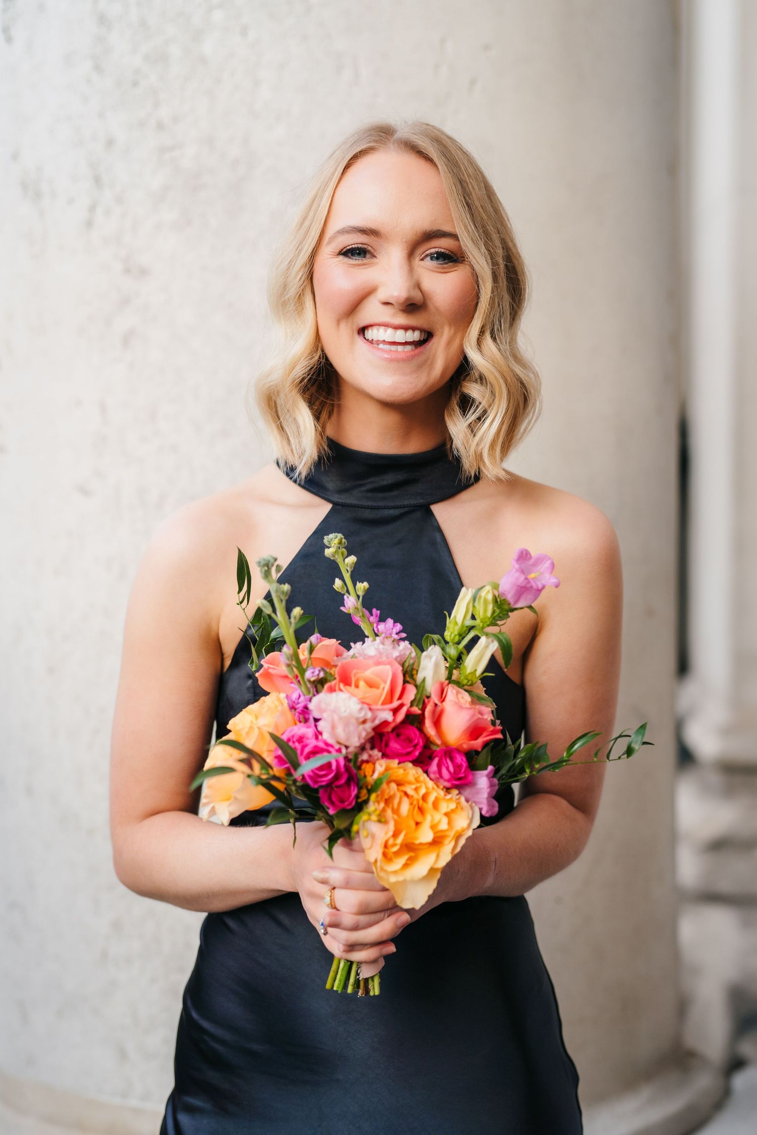 Bridesmaids in black dresses hold vibrant orange, yellow and pink floral bouquets against a light stone wall. City Hall Dublin Photographer