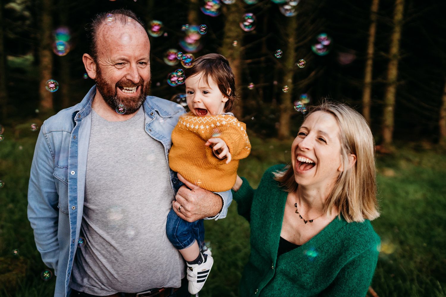 A cheerful family moment outdoors with soap bubbles floating in the forest as they laugh together in natural lighting. family PHOTOGRAPHER D