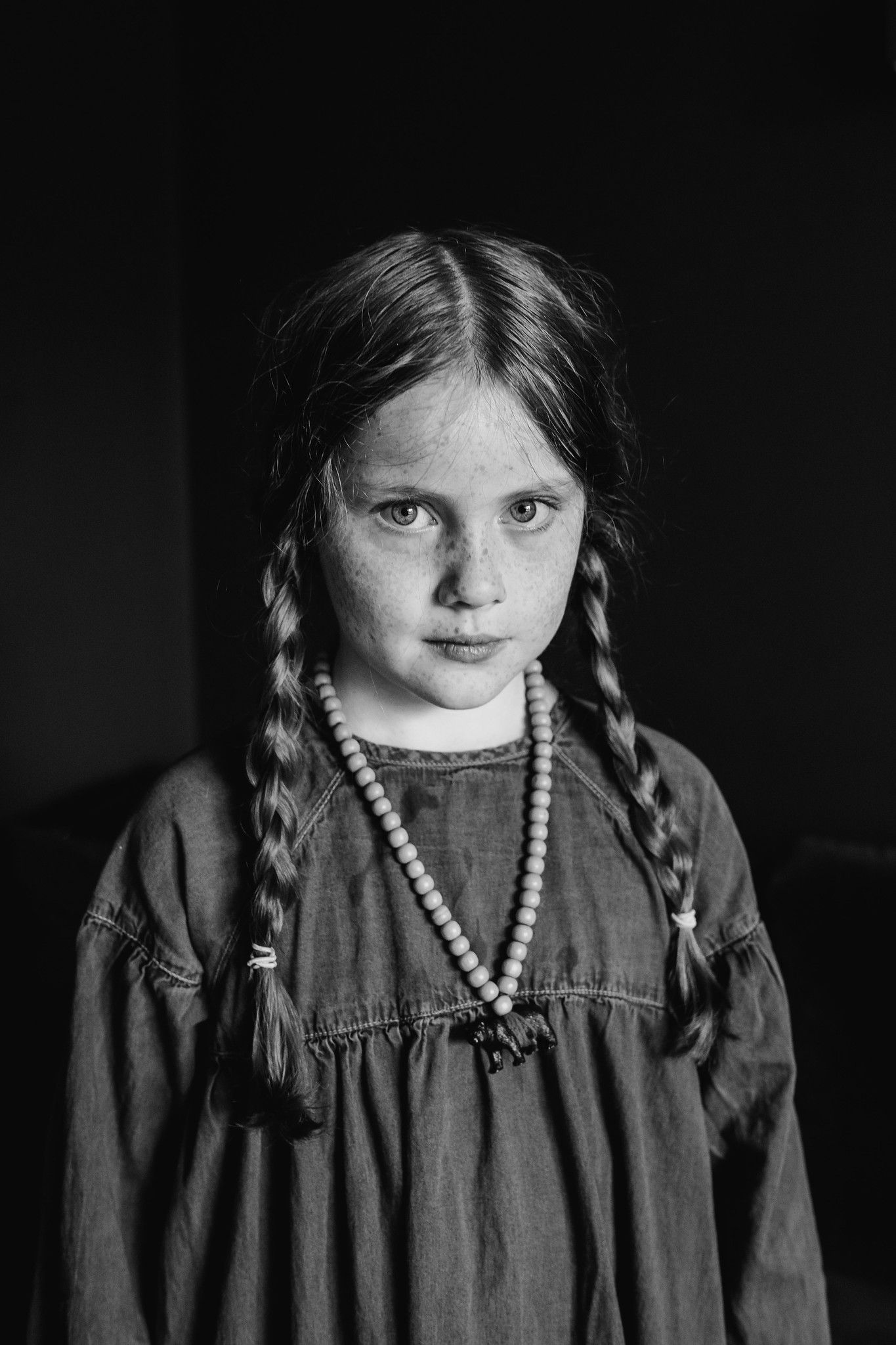 Black and white portrait of a young child wearing braids, a beaded necklace, and a dark dress against a dark background. PHOTOGRAPHER Dublin