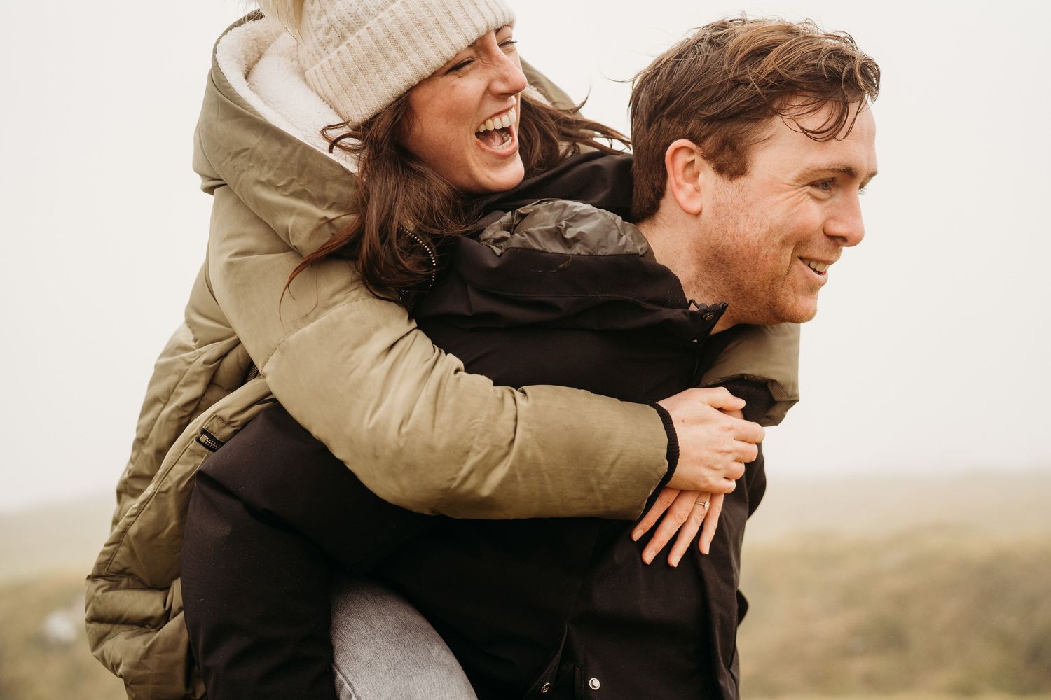 A couple sharing a playful piggyback ride outdoors in winter clothing against a misty natural landscape. PHOTOGRAPHER Dublin ireland