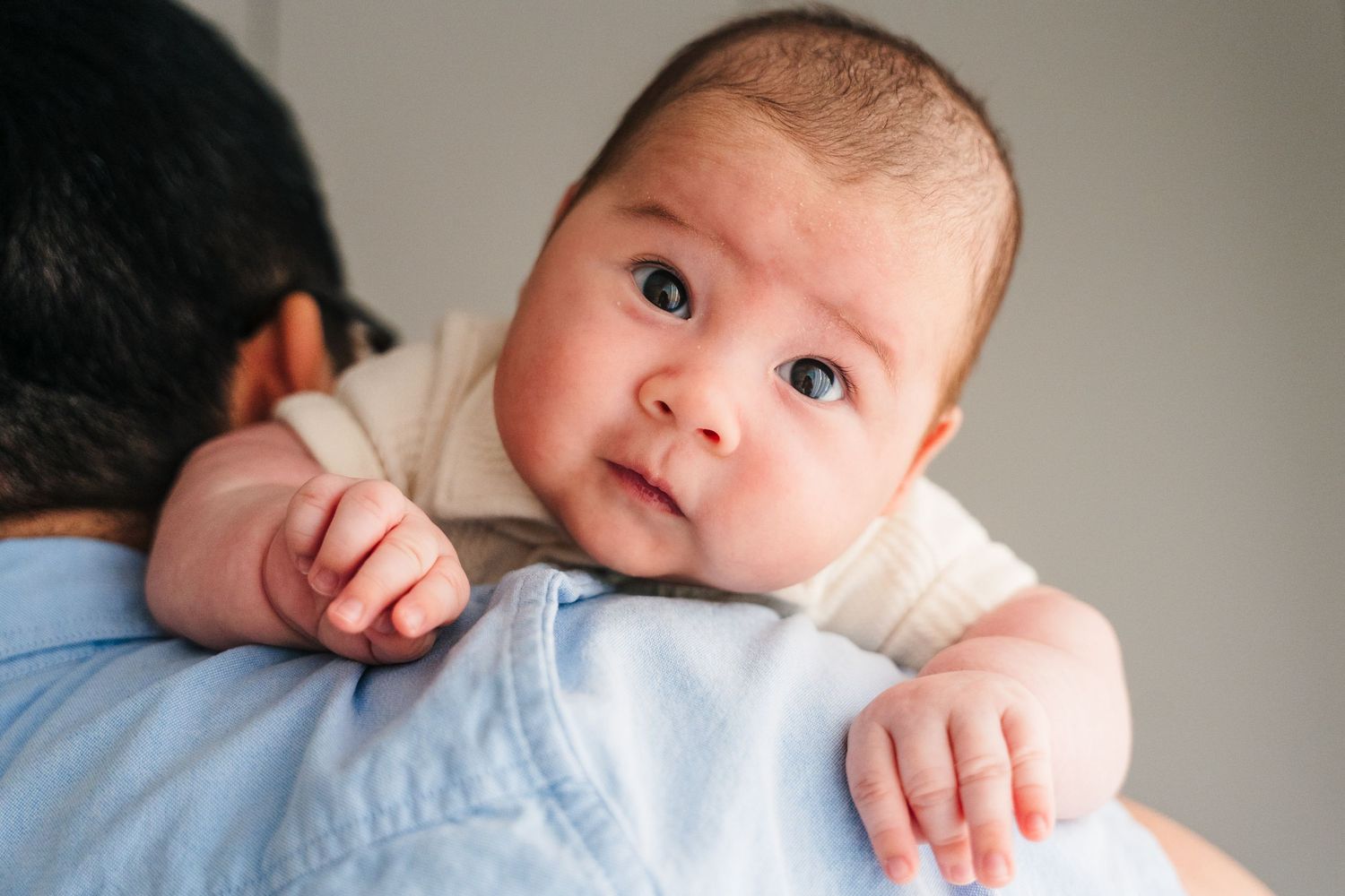 Newborn baby with watchful eyes wearing light colored clothing rests on patterned bedding while being comforted. family PHOTOGRAPHER Dublin