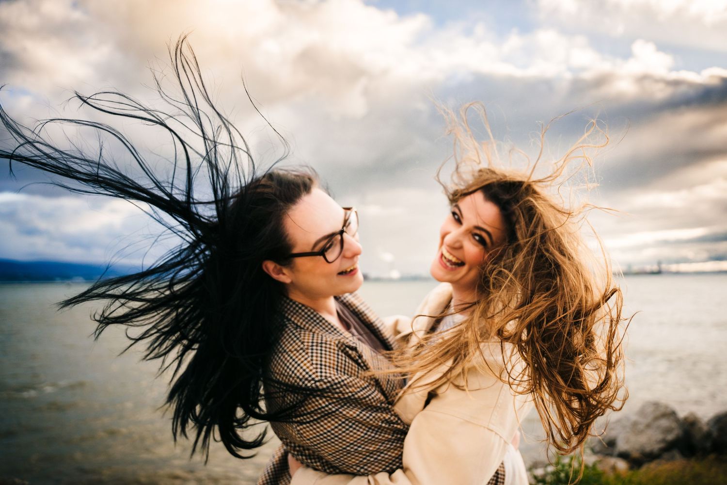 Two friends with windswept hair laugh joyfully together against a dramatic cloudy beach backdrop at sunset. PHOTOGRAPHER Dublin ireland