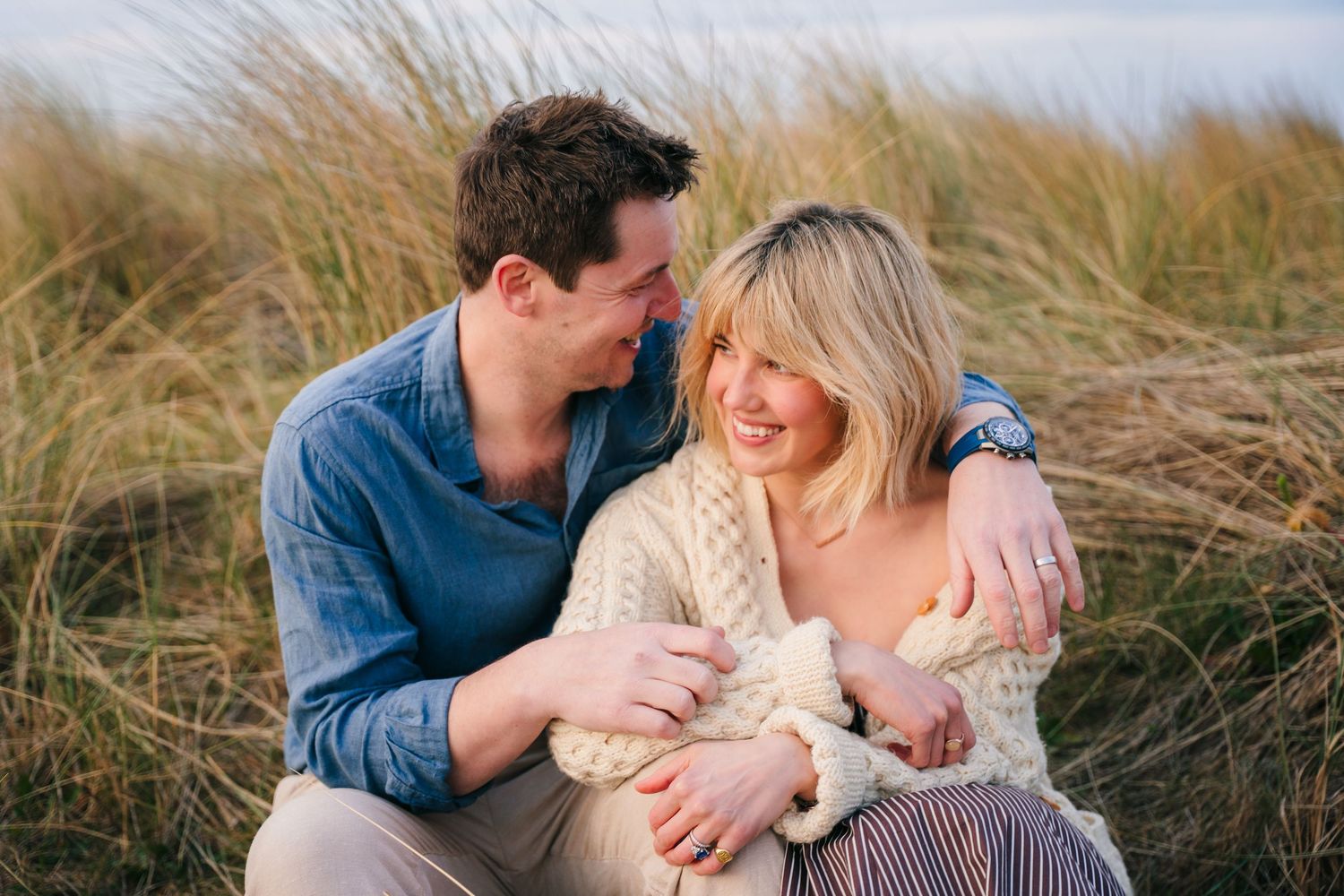 A loving couple embraces while sitting together in tall grass during a golden hour photoshoot at the beach. PHOTOGRAPHER Dublin ireland