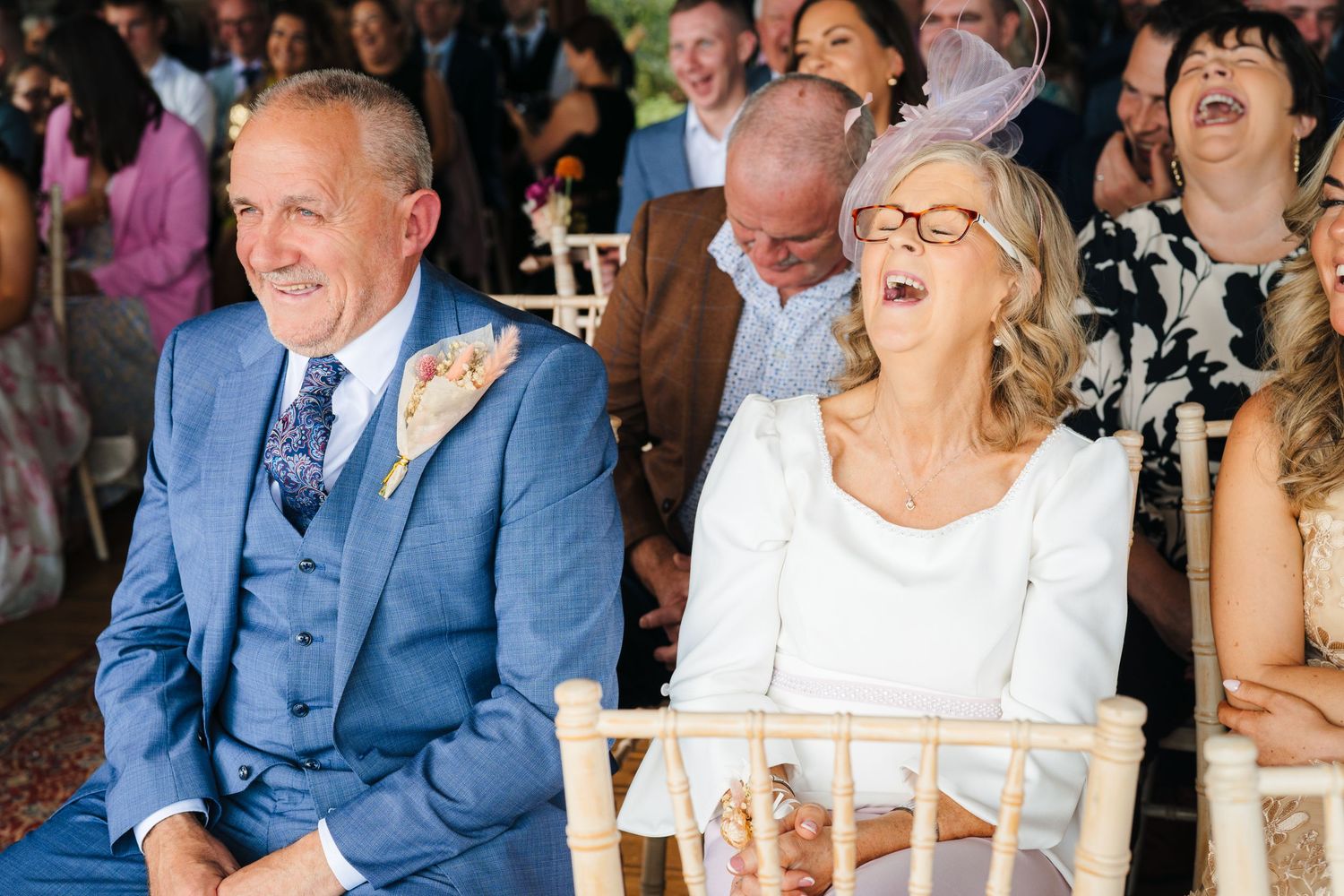 Wedding guests laughing and enjoying themselves during an outdoor ceremony with white chairs. LGBTQ Ashley Park Wedding Photographer