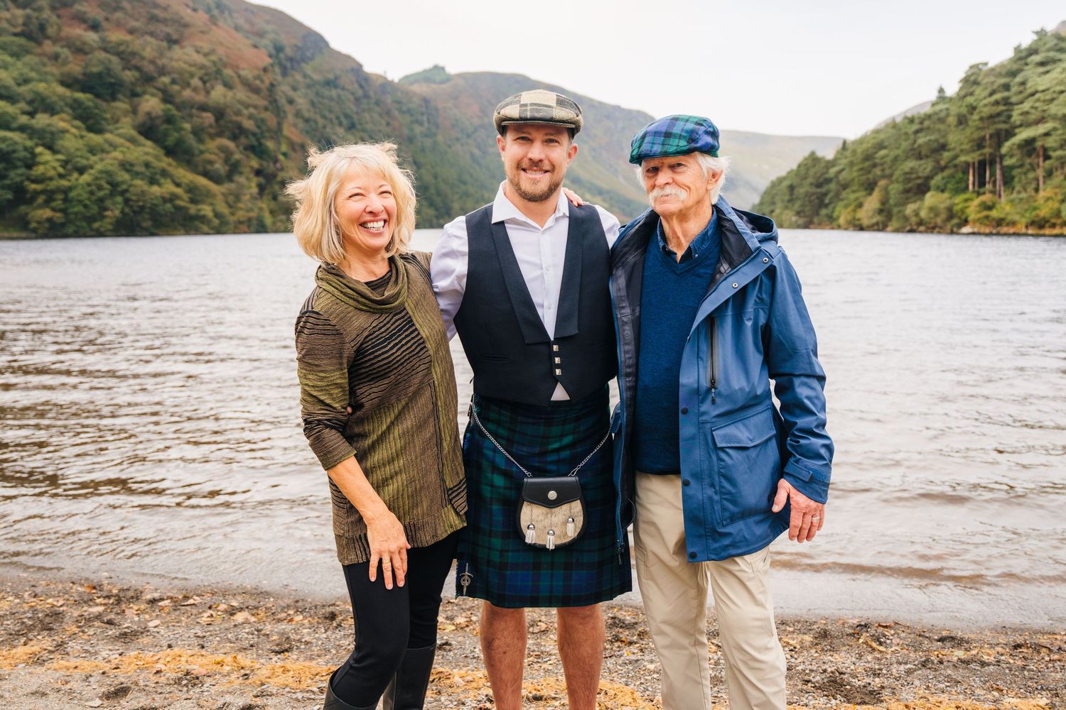 A group poses together on a rocky beach with mountains and a lake in the background.Family photogrAPHER DUBLIN