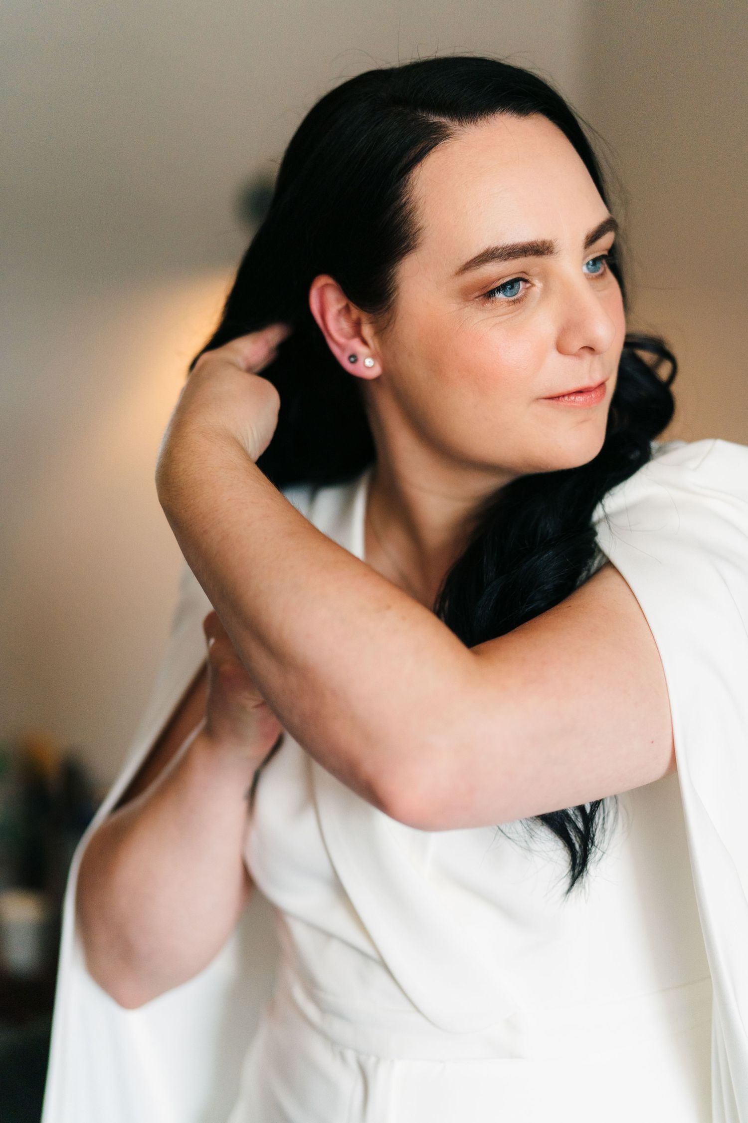Woman in white attire runs fingers through dark hair while looking to the side in a soft-lit setting. House Dublin Wedding Photographer