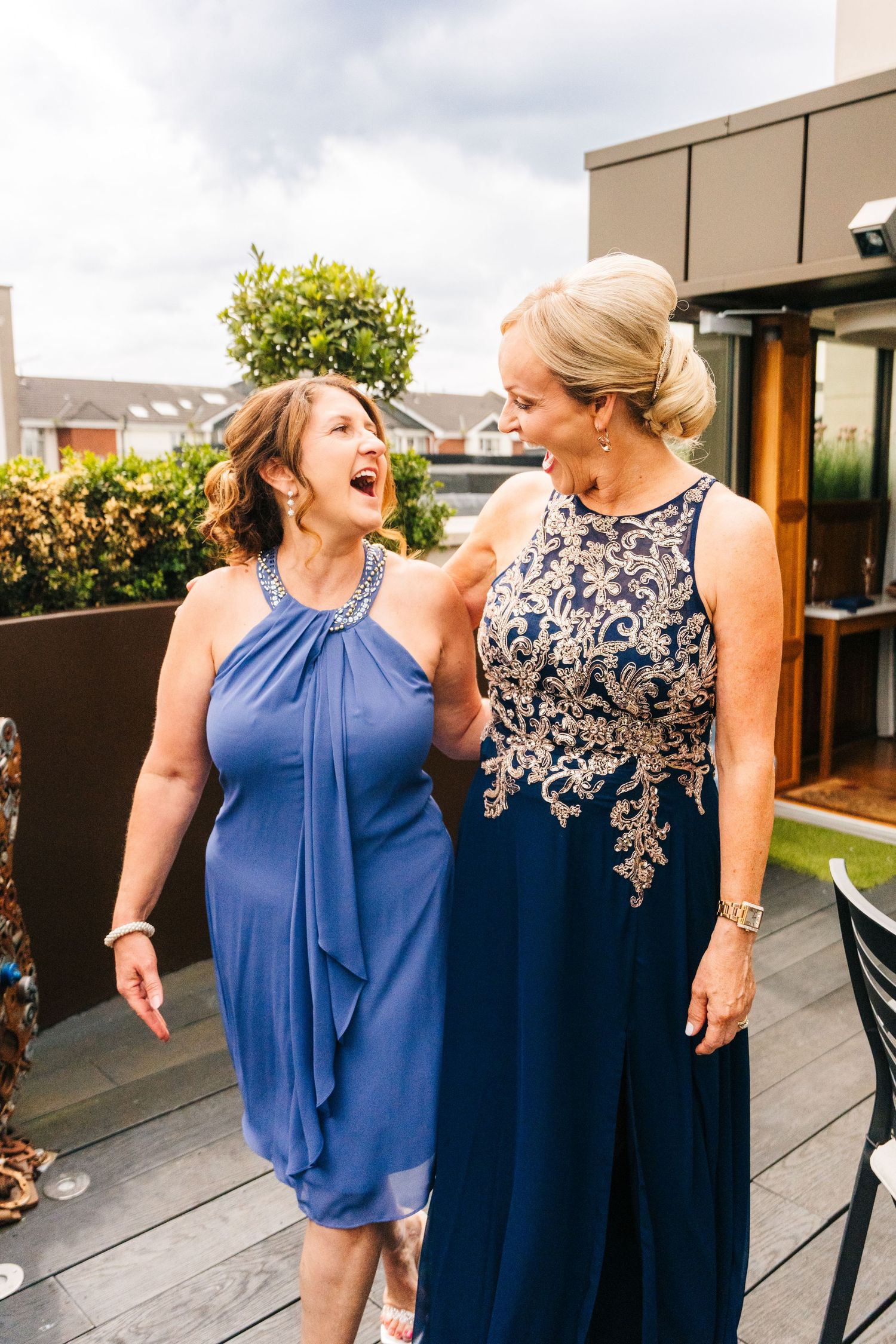 Two women in elegant blue formal dresses share a joyful laugh together on an outdoor deck. PHOTOGRAPHER Dublin Ireland Wedding