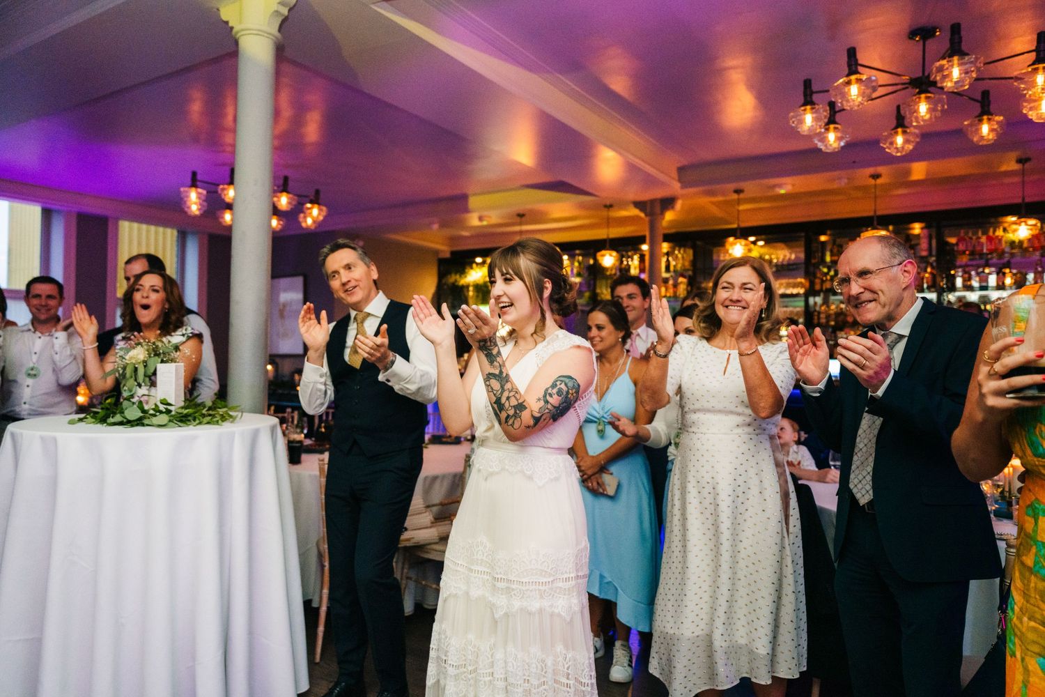 A wedding reception scene with purple lighting and guests celebrating at a white-clothed table. PHOTOGRAPHER Dublin Ireland Wedding