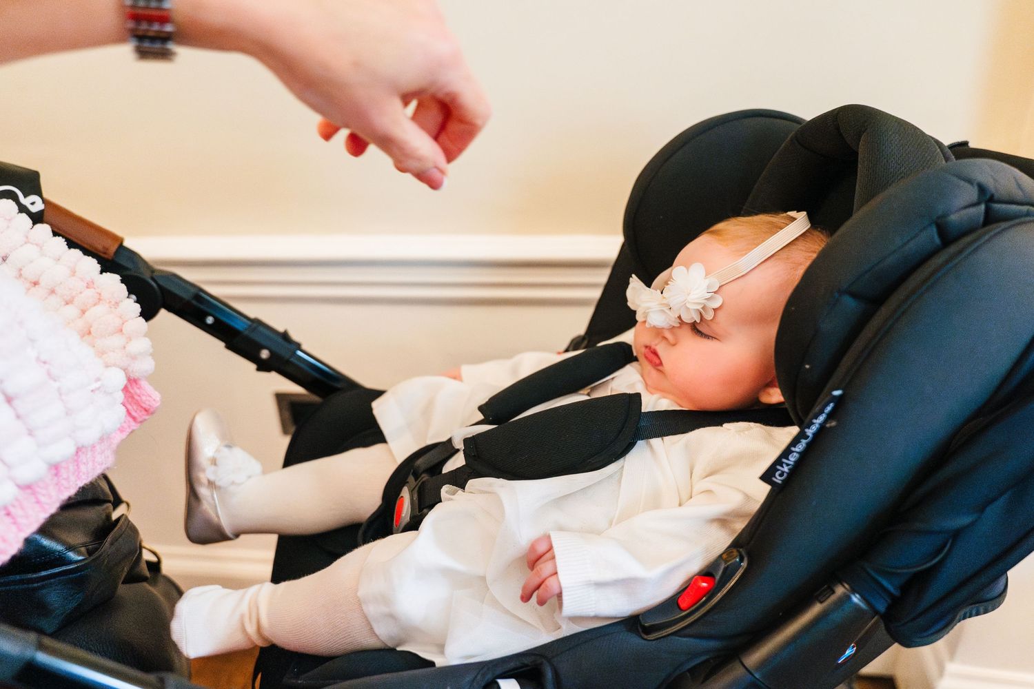 Baby sleeping in a navy blue car seat wearing a white outfit with a hand reaching to adjust the safety straps.  Wedding PHOTOGRAPHER Dublin