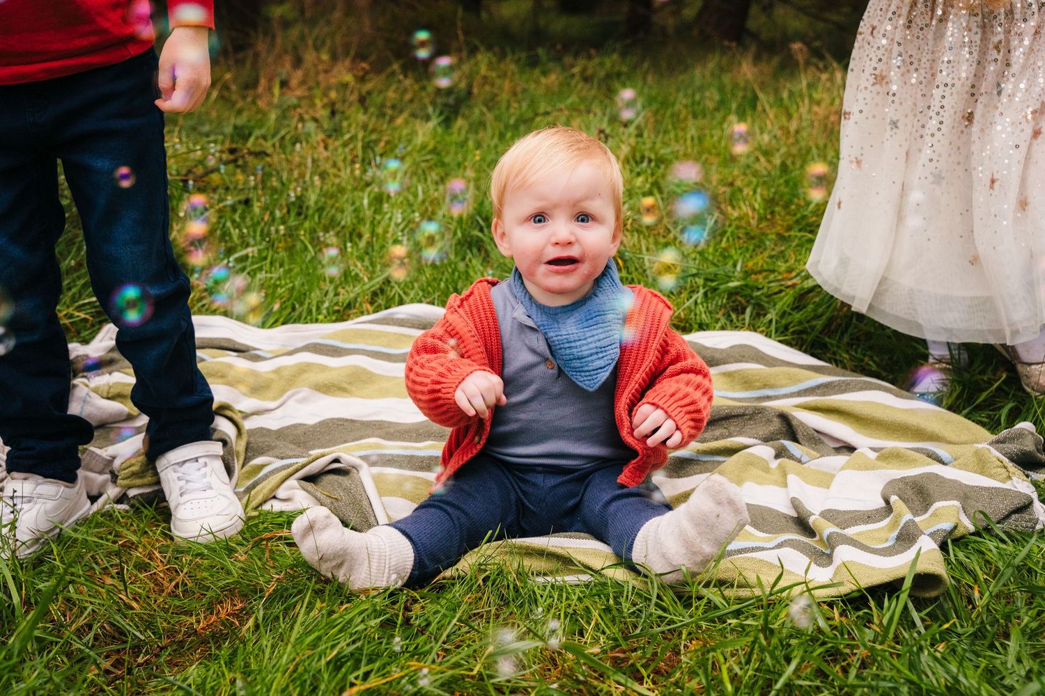 Baby wearing red and blue outfit sits on striped blanket in grass.