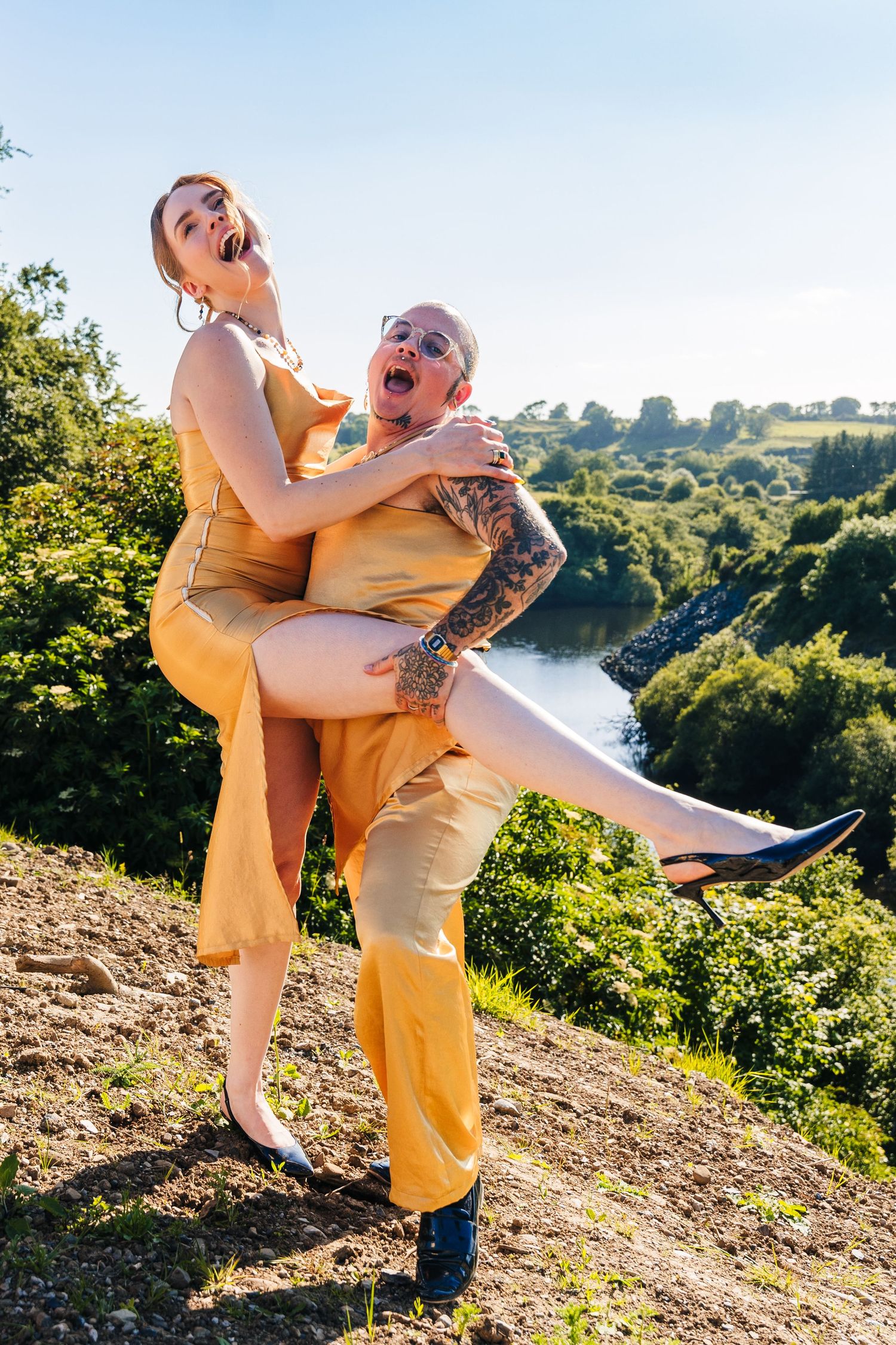 Poulaphouca Wedding photA joyful couple in matching yellow outfits laugh together while posing on a hillside overlooking a scenic landscape.