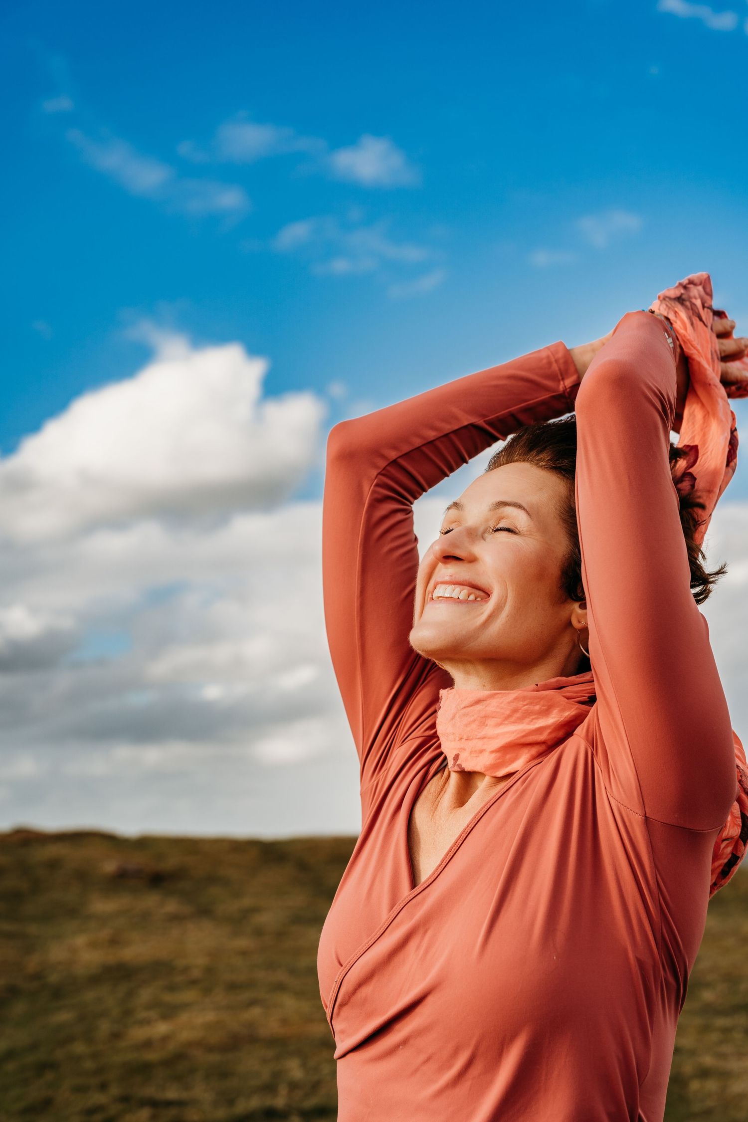 Person in coral-colored top with raised arms enjoying sunny outdoor moment against blue sky with clouds. Portrait PHOTOGRAPHER Dublin