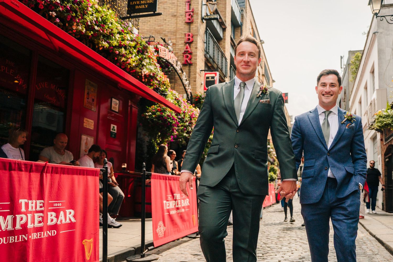 Two people in suits walk happily together past a red storefront with The Temple Bar signage in Dublin. Same sex wedding PHOTOGRAPHER Dublin