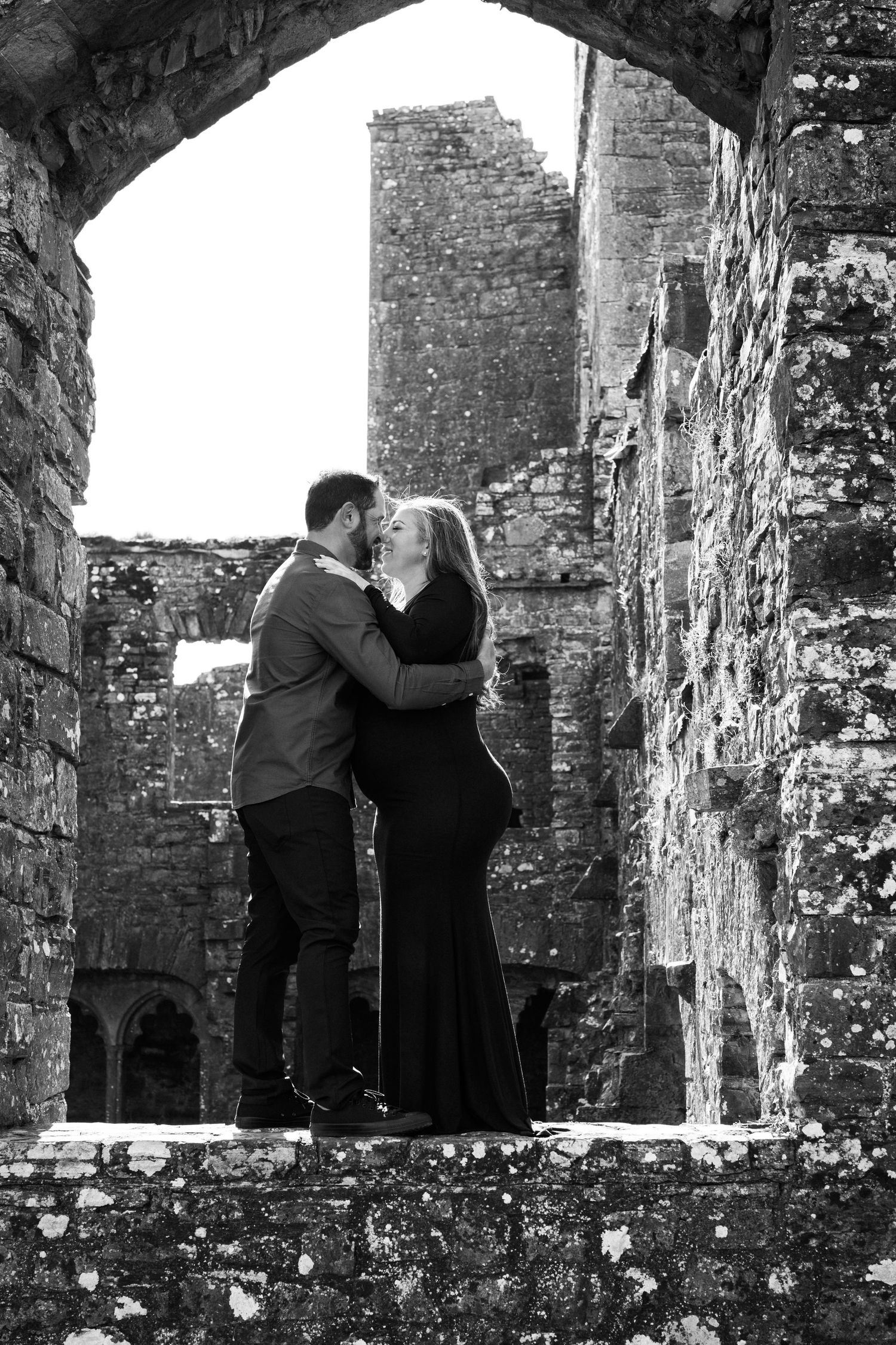 A romantic black and white photo of a couple embracing under a stone archway at an ancient castle ruin. Family PHOTOGRAPHER Dublin