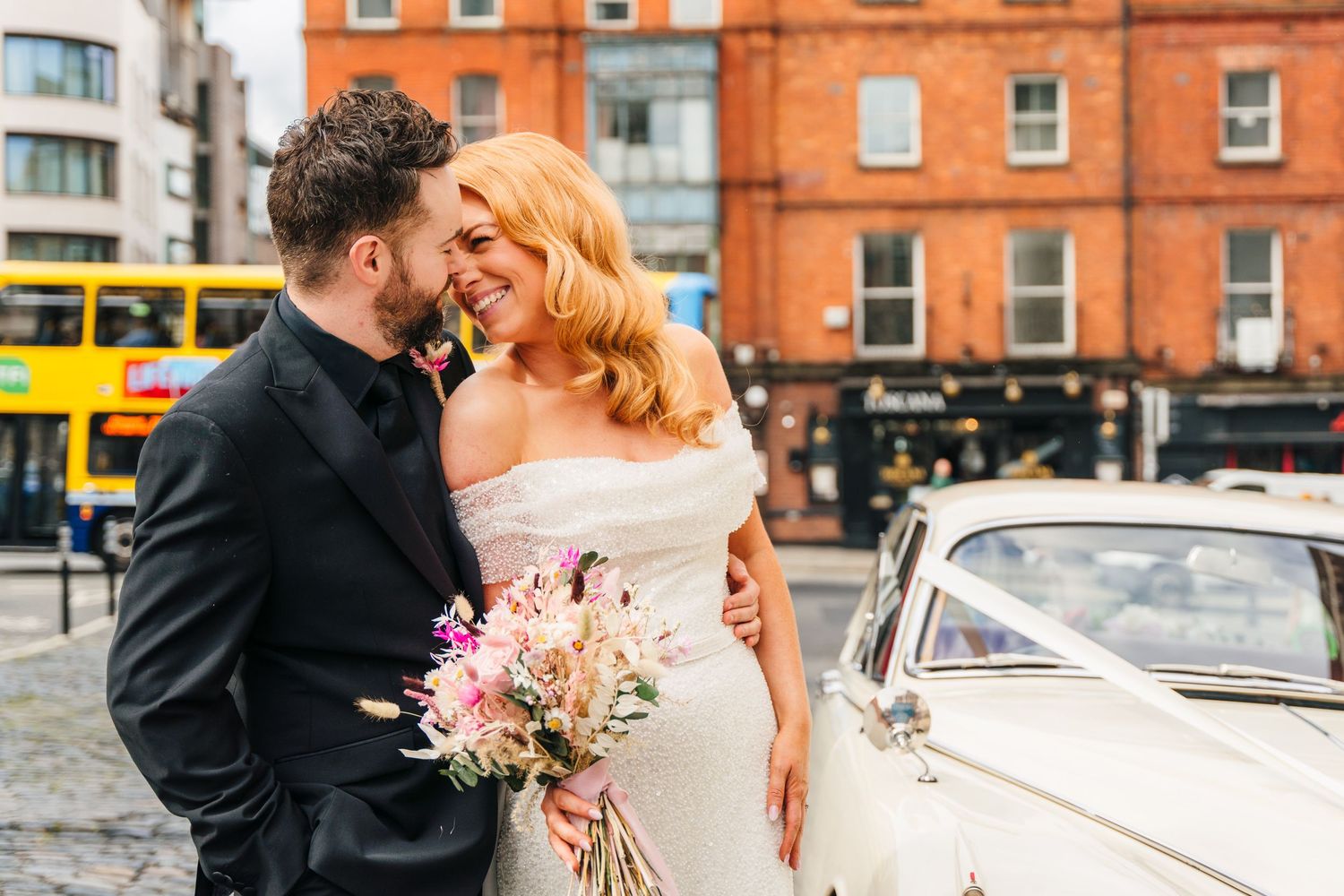 Wedding couple sharing a romantic moment near a vintage car with orange brick buildings in the background.  city hall dublin wedding photos