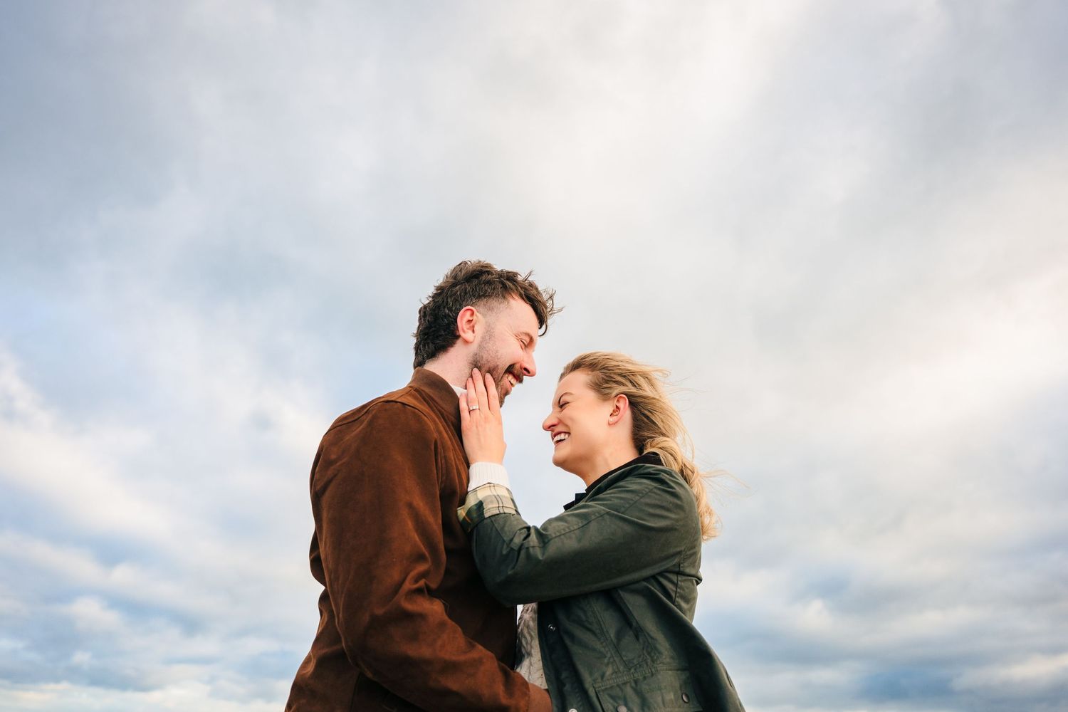 A romantic couple laughs together against a cloudy sky, with one wearing a brown jacket and the other in green. couple's photographer dublin