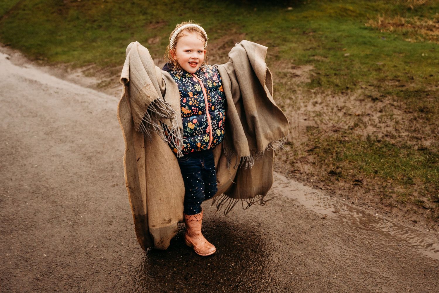 Child wearing an oversized brown coat with floral dress and brown boots stands on a pathway outdoors.nfamily PHOTOGRAPHER Dublin