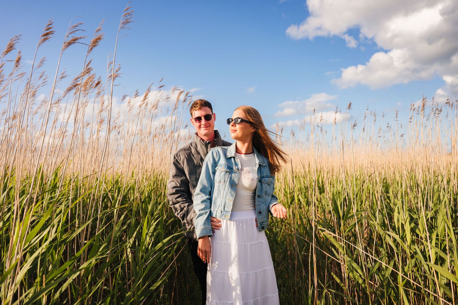 Couple walking through tall golden grass against blue sky with white clouds during outdoor photo session. PHOTOGRAPHER Dublin ireland