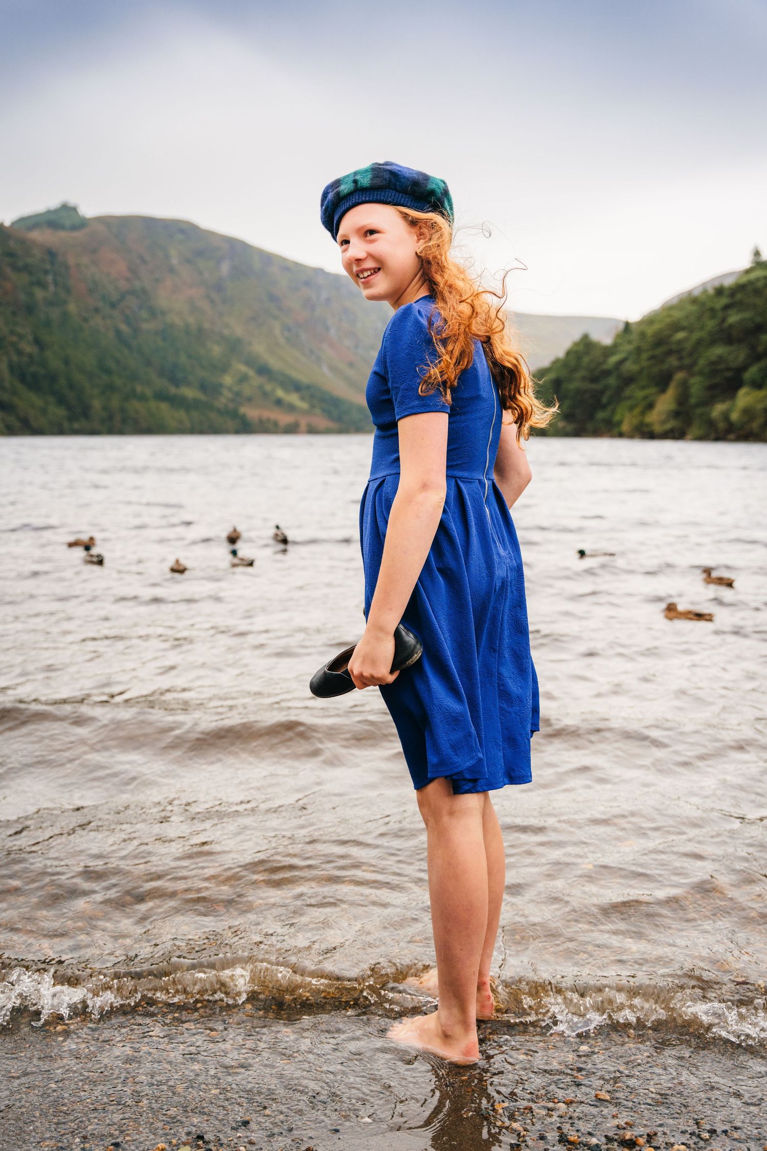 Young child wearing a blue dress plays at the edge of a lake with mountains in the background. Family photogrAPHER DUBLIN