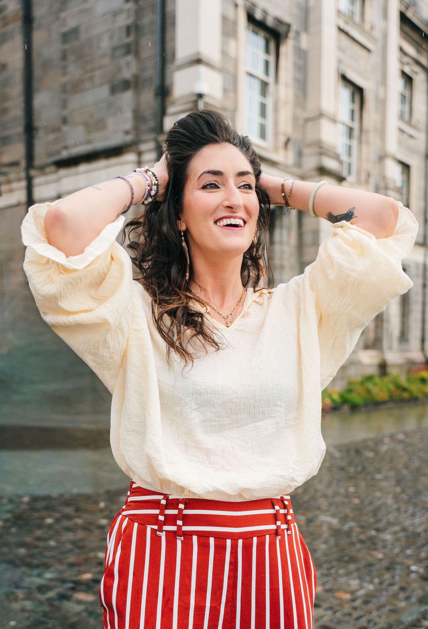Fashion model wearing cream sweater and red striped skirt poses joyfully against stone building backdrop. professional headshots dublin