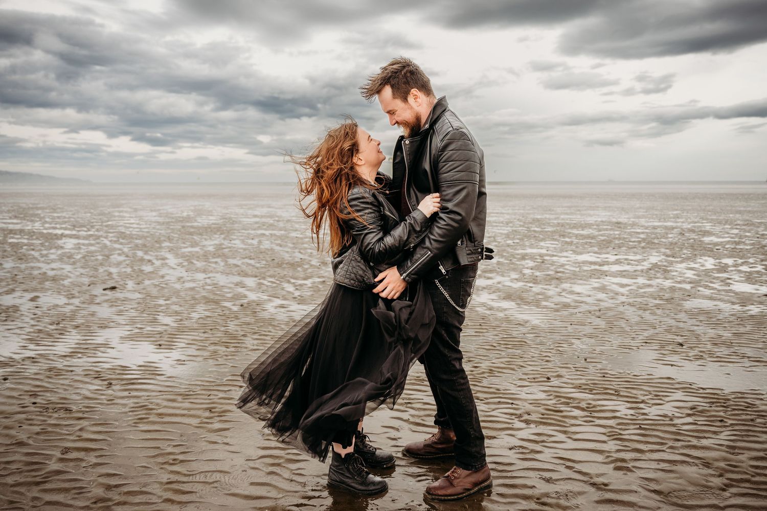 Couple shares intimate moment on windswept beach under moody grey clouds. engagement PHOTOGRAPHER Dublin