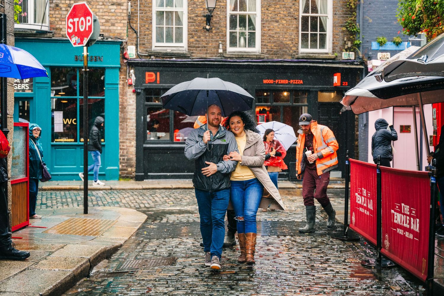 Pedestrians walking with umbrellas on a rainy cobblestone street with red market stalls and brick buildings. Proposal Dublin Photographer