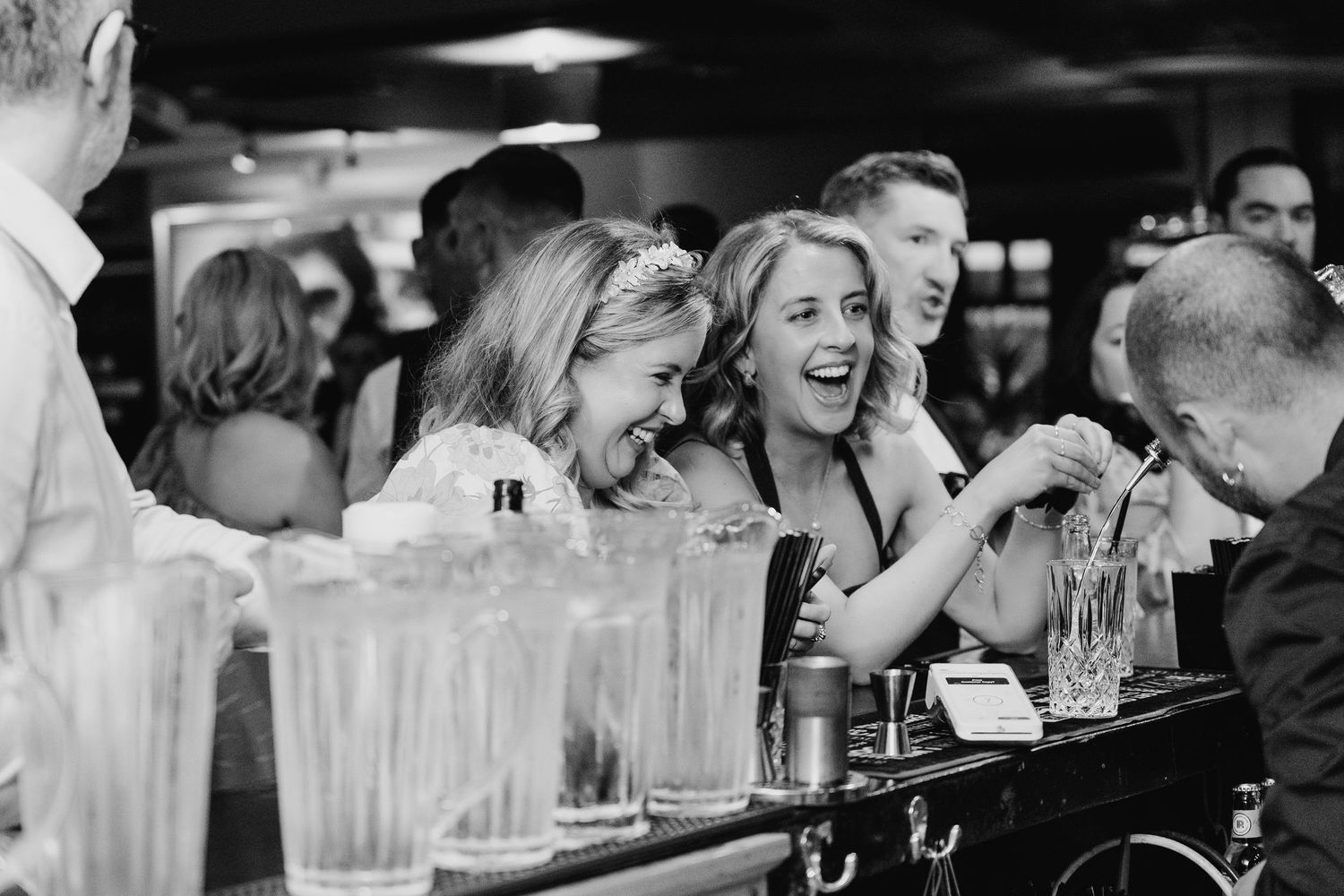 Wedding guests enjoy drinks and laughter at a dimly lit bar counter. Marco Pierre Dublin Wedding Photographer