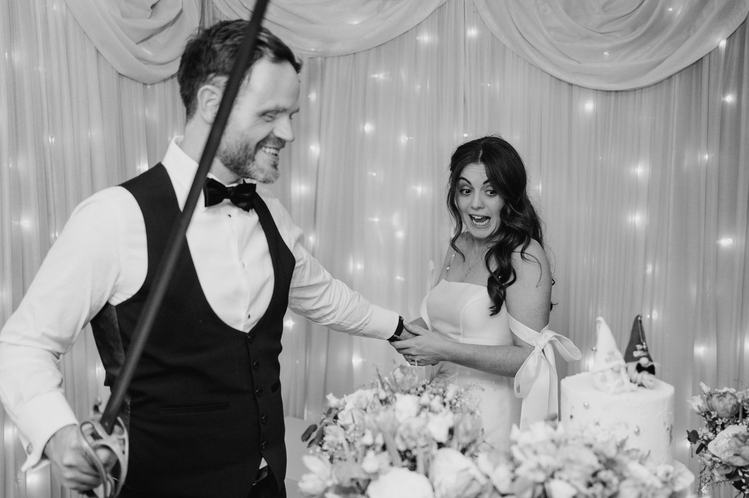Castle Leslie Wedding Photographer Dublin Black and white photo of newlyweds cutting cake together at reception with fairy light backdrop.