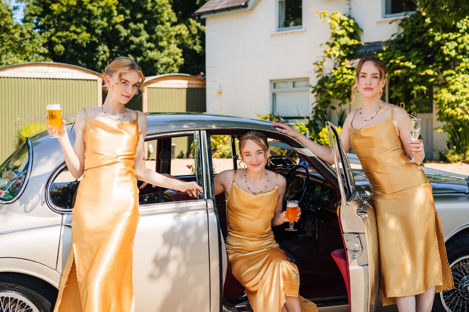 Poulaphouca Wedding photographer wicklow  A classic vintage car parked outdoors with ladies in gold satin dresses standing around it.