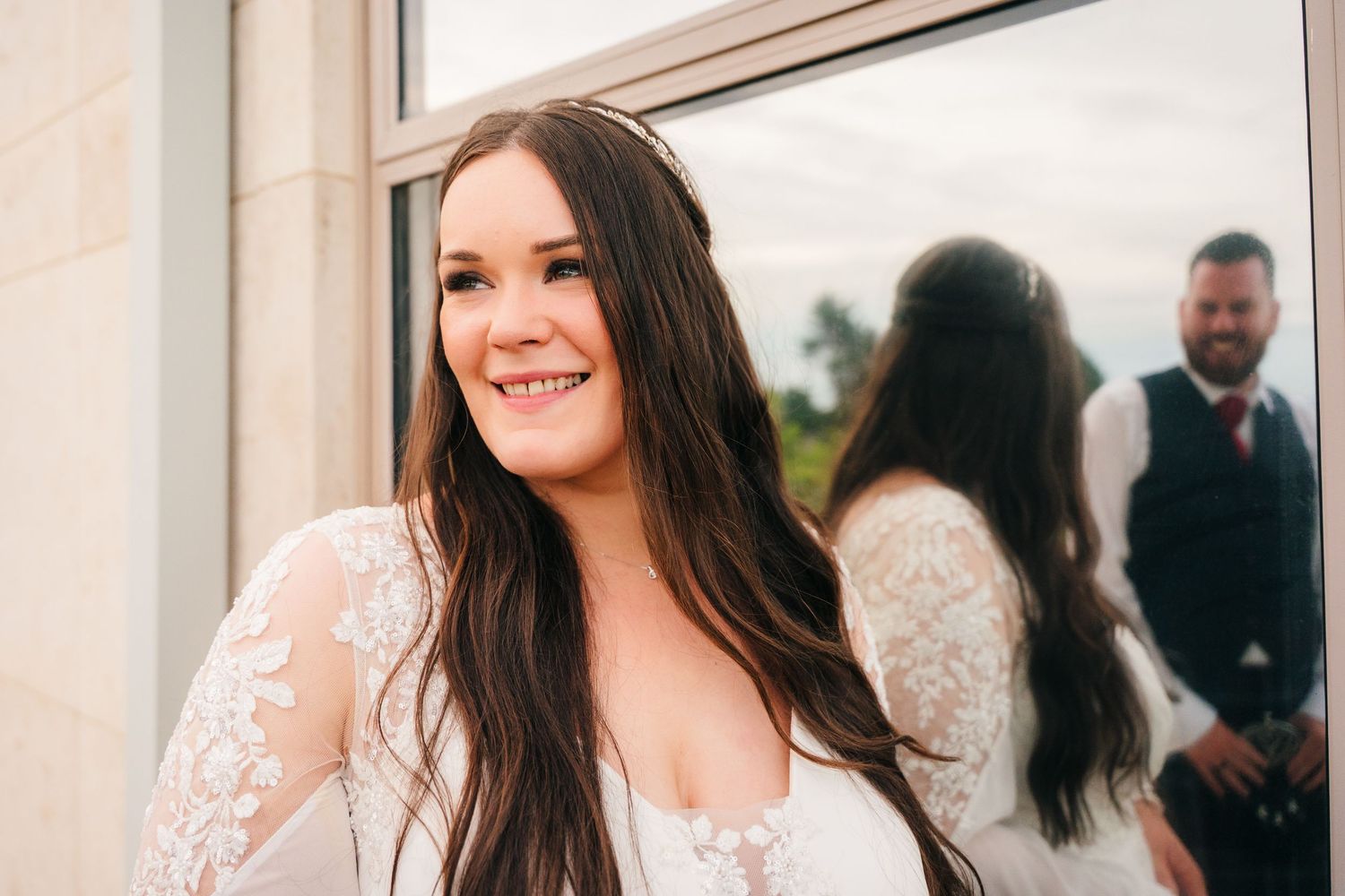 A joyful bride in lace sleeves poses while her reflection shows in a window. Portmarnock Hotel Wedding PHOTOGRAPHER Dublin