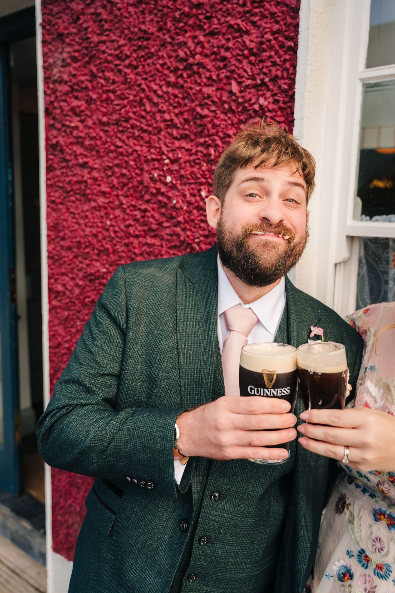 Person in a gray suit holding two dark pints of Guinness beer against a vibrant red ivy-covered wall. . Village Barn Tyrrellspass wedding ph